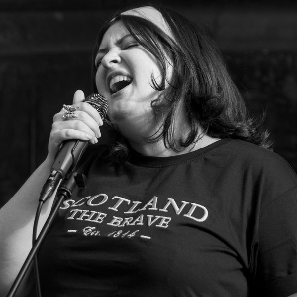 Black-and-white close-up of a woman singing into a wired microphone, wearing a T-shirt printed “Scotland the Brave.”