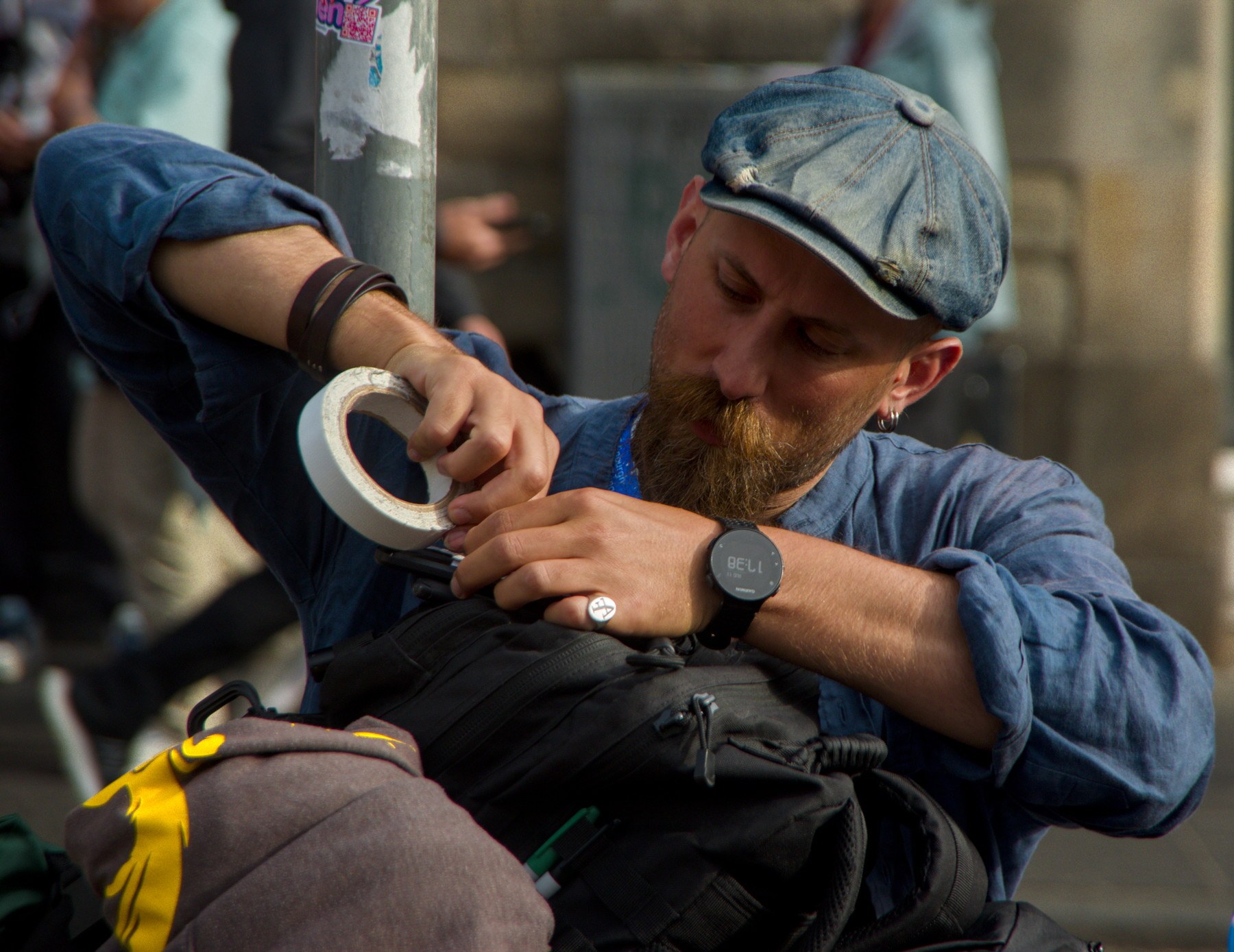 Bearded man in a blue flat cap and rolled-sleeve shirt using a roll of tape to secure a black bag on a city street.