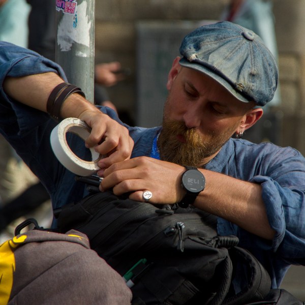 Bearded man in a blue flat cap and rolled-sleeve shirt using a roll of tape to secure a black bag on a city street.