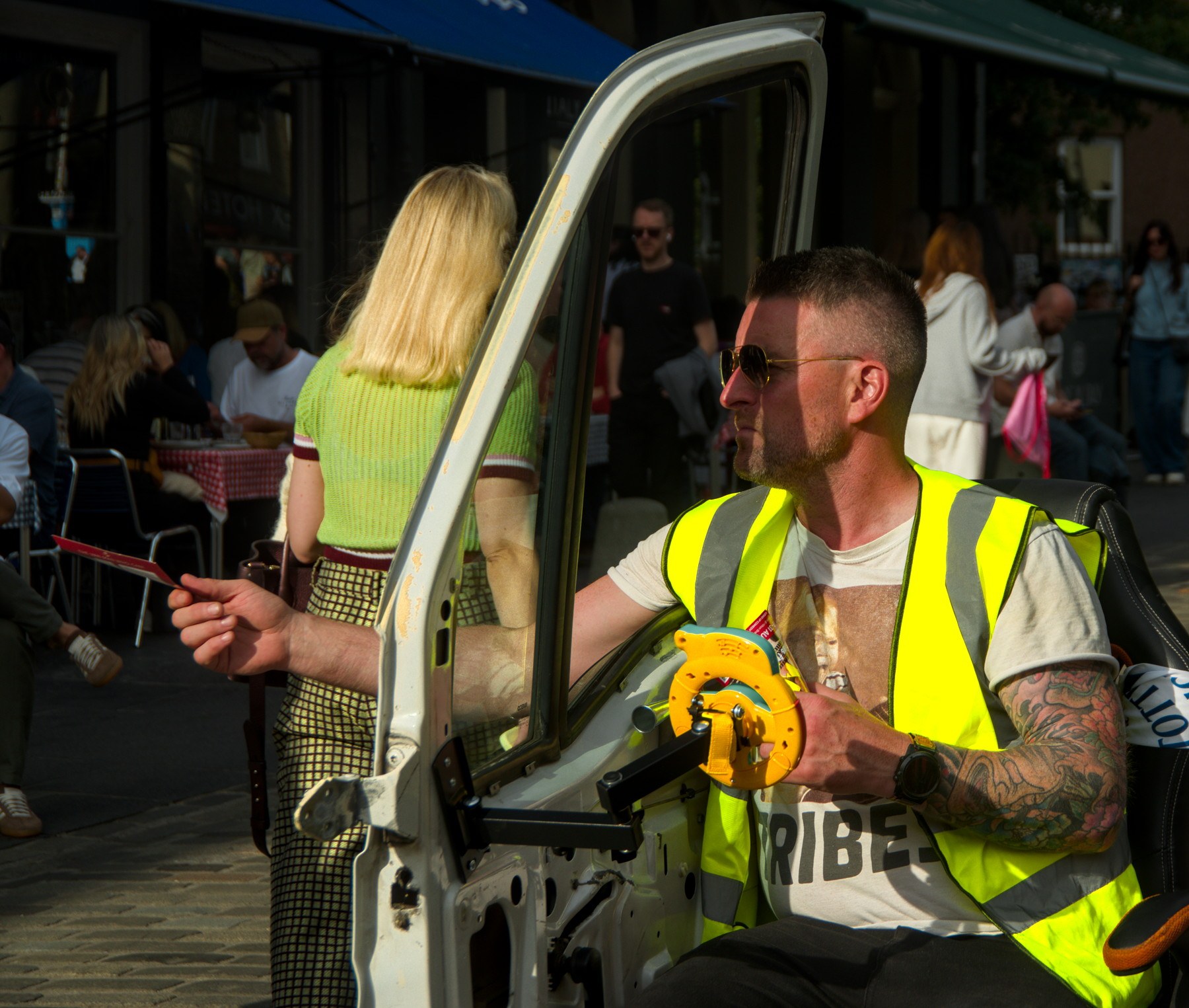 Tattooed man in a fluorescent yellow safety vest sits in an open vehicle doorway, extending a thin rod toward a busy Edinburgh pavement café.