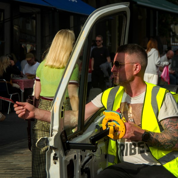Tattooed man in a fluorescent yellow safety vest sits in an open vehicle doorway, extending a thin rod toward a busy Edinburgh pavement café.