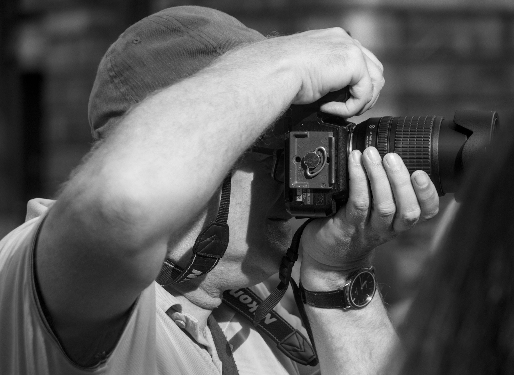 Close-up black-and-white view of a man in a cap raising a camera with a long lens to photograph something beyond the frame.