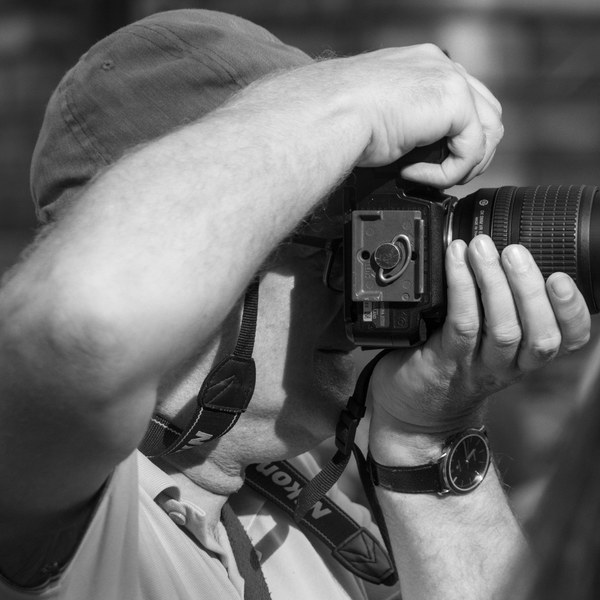 Close-up black-and-white view of a man in a cap raising a camera with a long lens to photograph something beyond the frame.