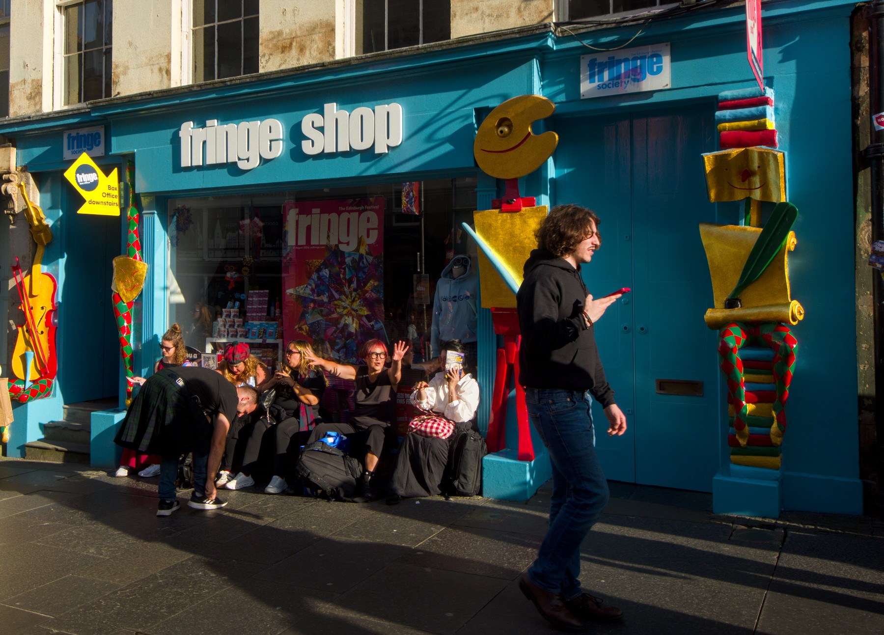 People sit and wave outside the turquoise Fringe Shop storefront as a man walks past looking at his phone.