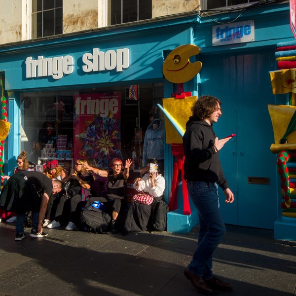 People sit and wave outside the turquoise Fringe Shop storefront as a man walks past looking at his phone.