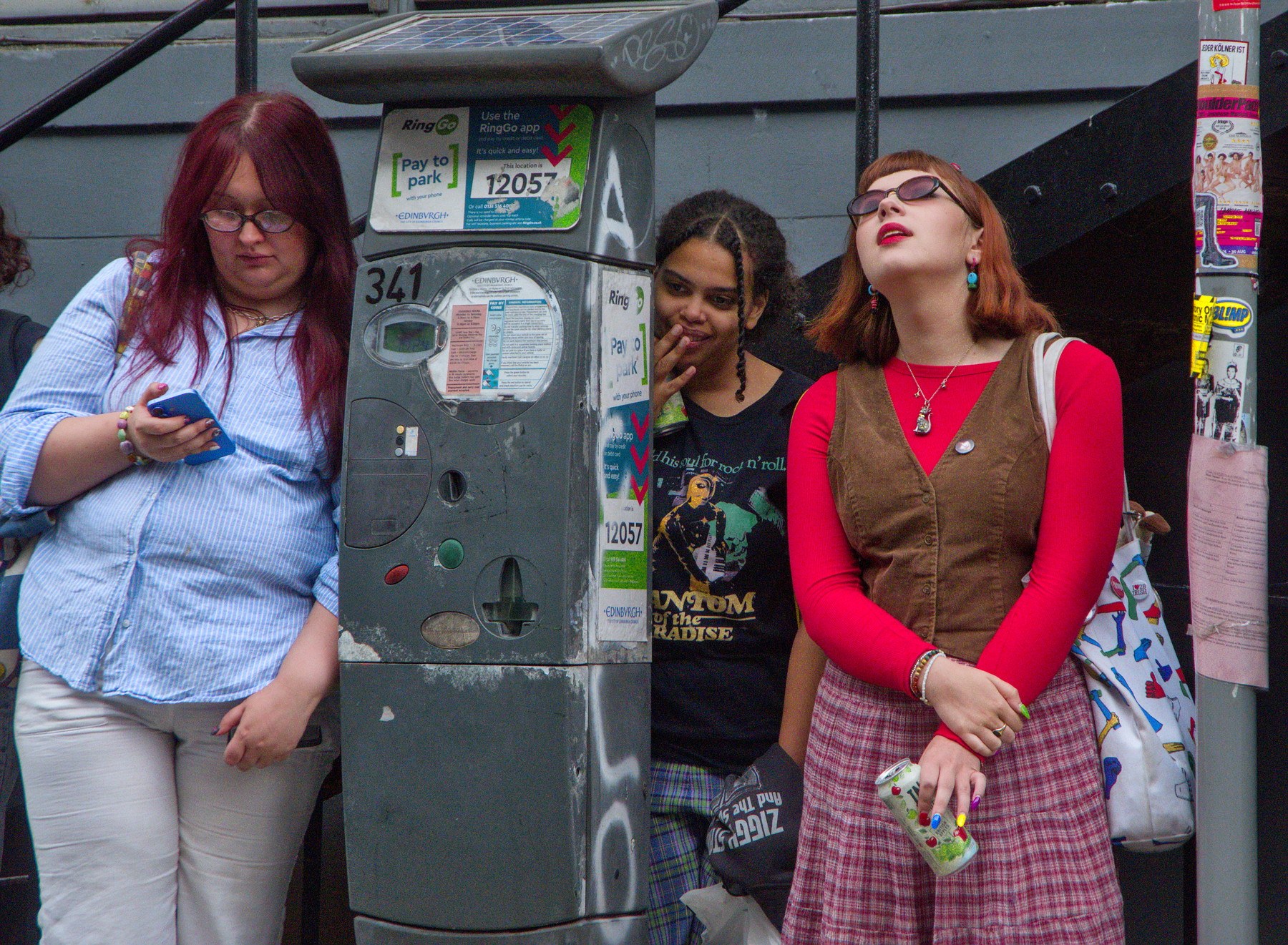 Three young women stand beside a sticker-covered parking meter. One looks at a blue phone, another peeks from behind the meter with her hand near her mouth, and a third in a red top and plaid skirt tilts her head back.