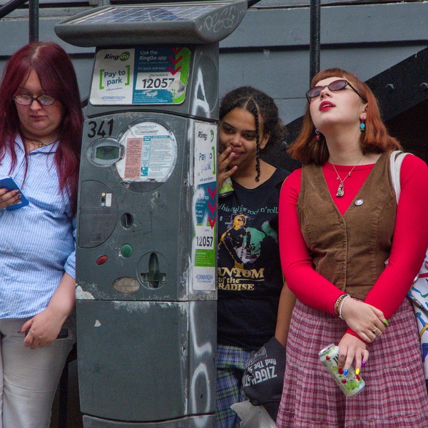 Three young women stand beside a sticker-covered parking meter. One looks at a blue phone, another peeks from behind the meter with her hand near her mouth, and a third in a red top and plaid skirt tilts her head back.