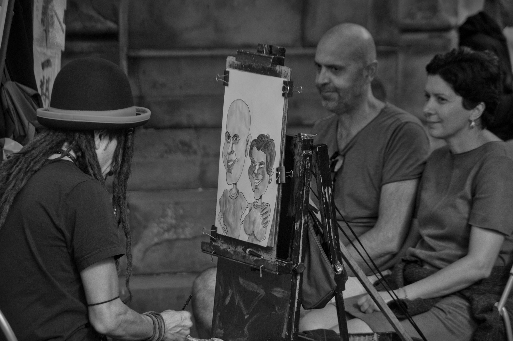 Street caricature artist in a bowler hat drawing a seated couple on an easel while they watch.