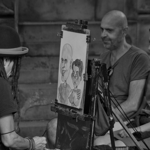 Street caricature artist in a bowler hat drawing a seated couple on an easel while they watch.
