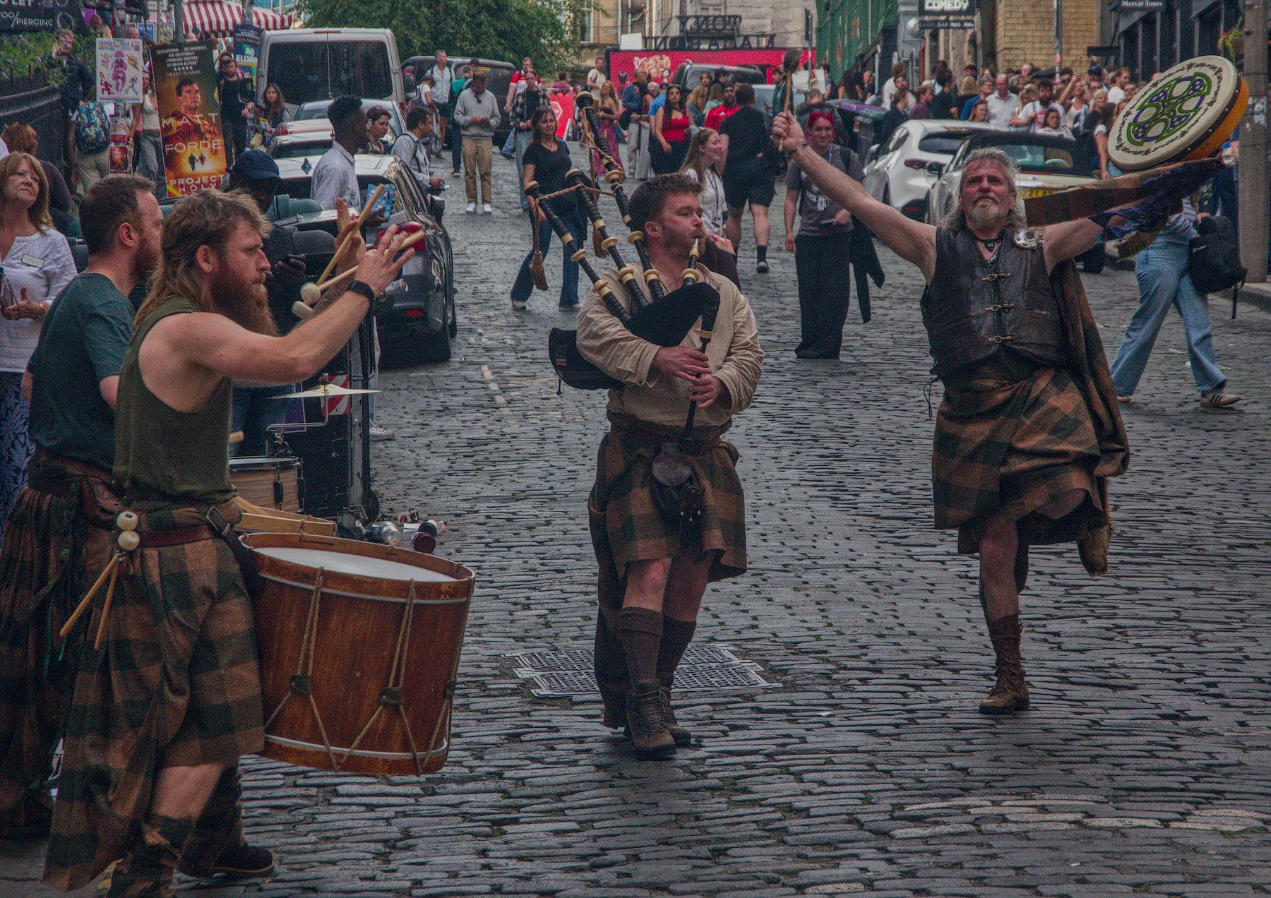 Three kilted street performers play a drum and bagpipes on a cobbled Edinburgh street as a man raises a painted round shield; festival-goers watch behind them.