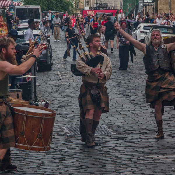 Three kilted street performers play a drum and bagpipes on a cobbled Edinburgh street as a man raises a painted round shield; festival-goers watch behind them.