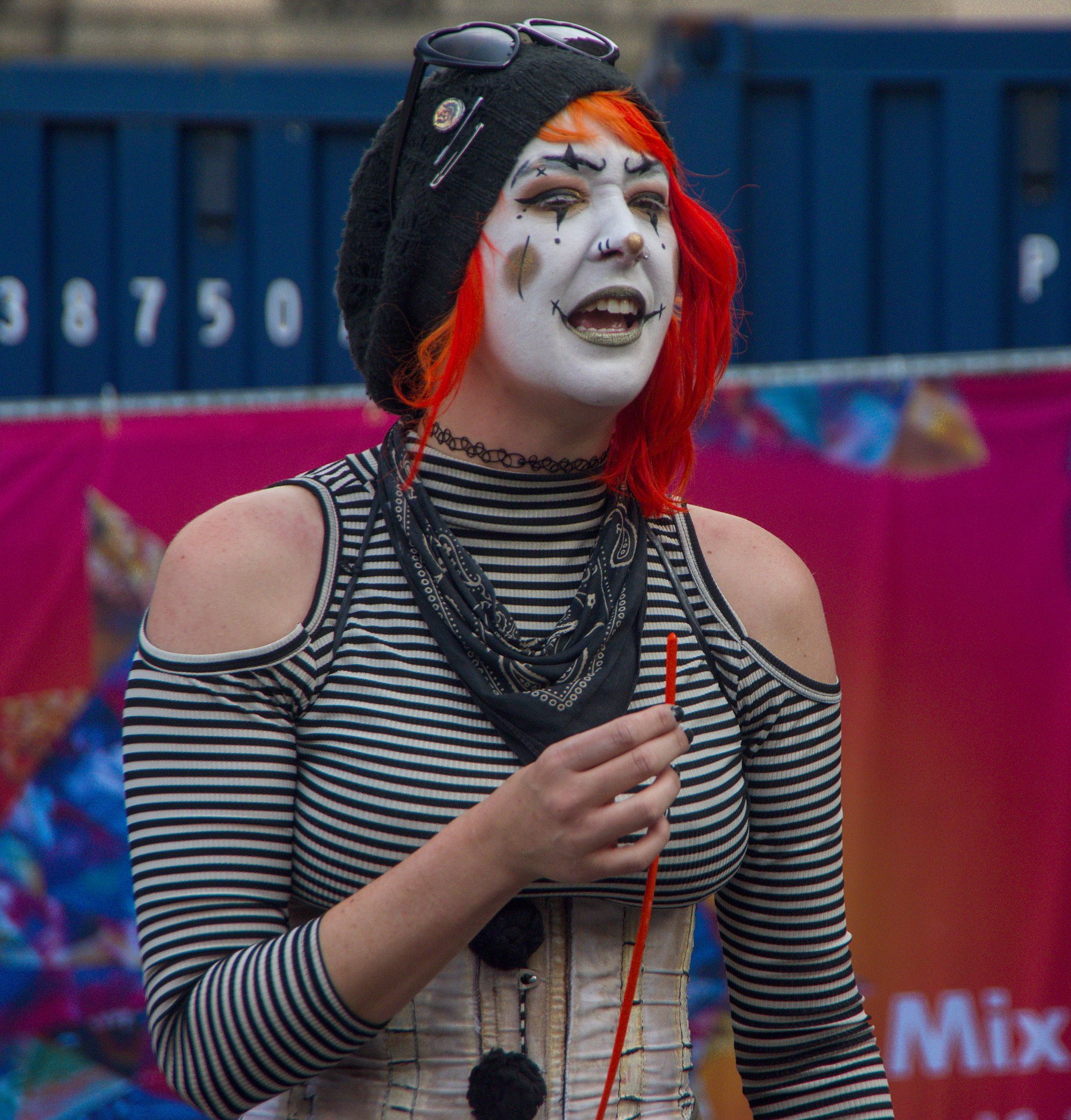 Festival perforrmer with white-and-black face paint, red hair and a striped top holds a red cord, mouth open, in front of blue barriers and a pink stall backdrop.