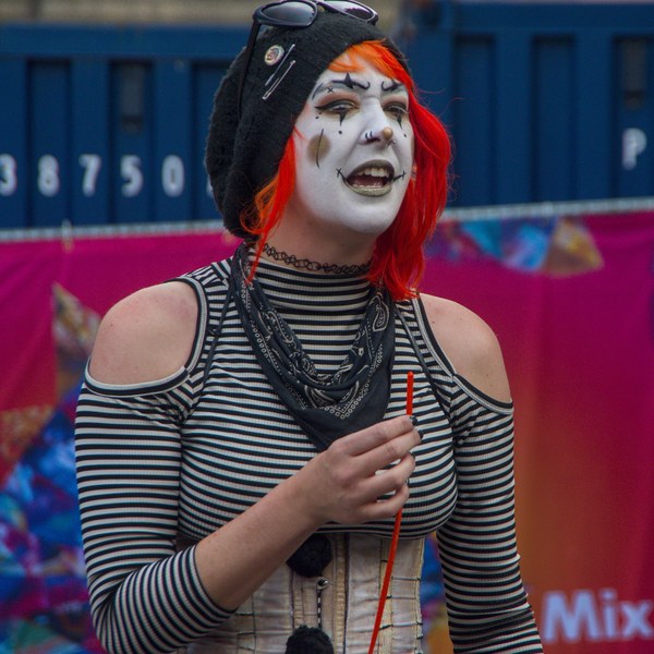 Festival perforrmer with white-and-black face paint, red hair and a striped top holds a red cord, mouth open, in front of blue barriers and a pink stall backdrop.