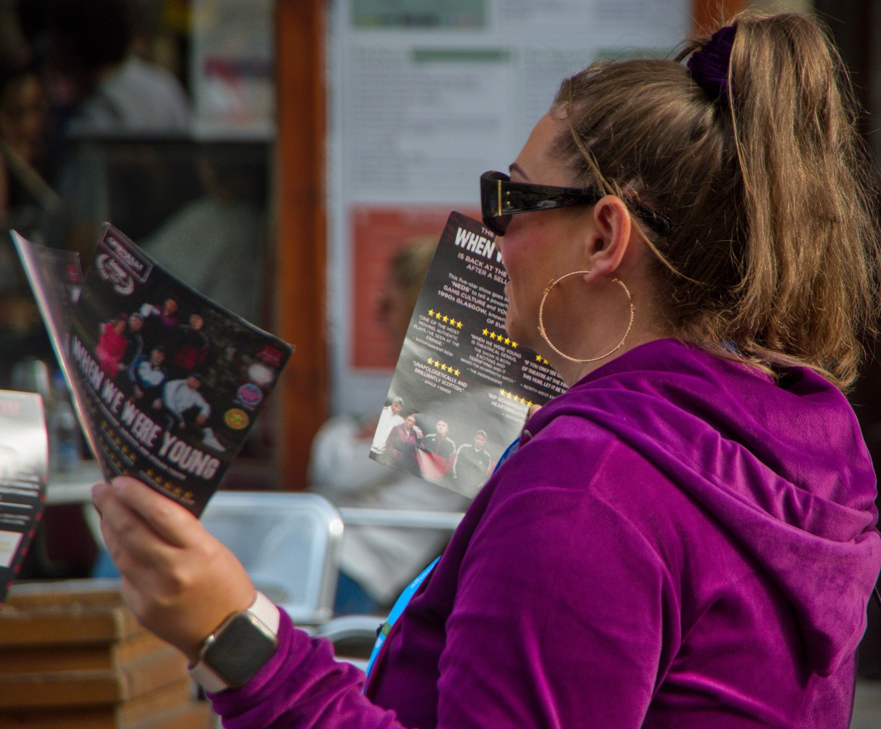 Woman in sunglasses and a purple hoodie reading a “When We Were Young” festival flyer.