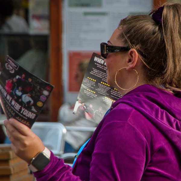 Woman in sunglasses and a purple hoodie reading a “When We Were Young” festival flyer.
