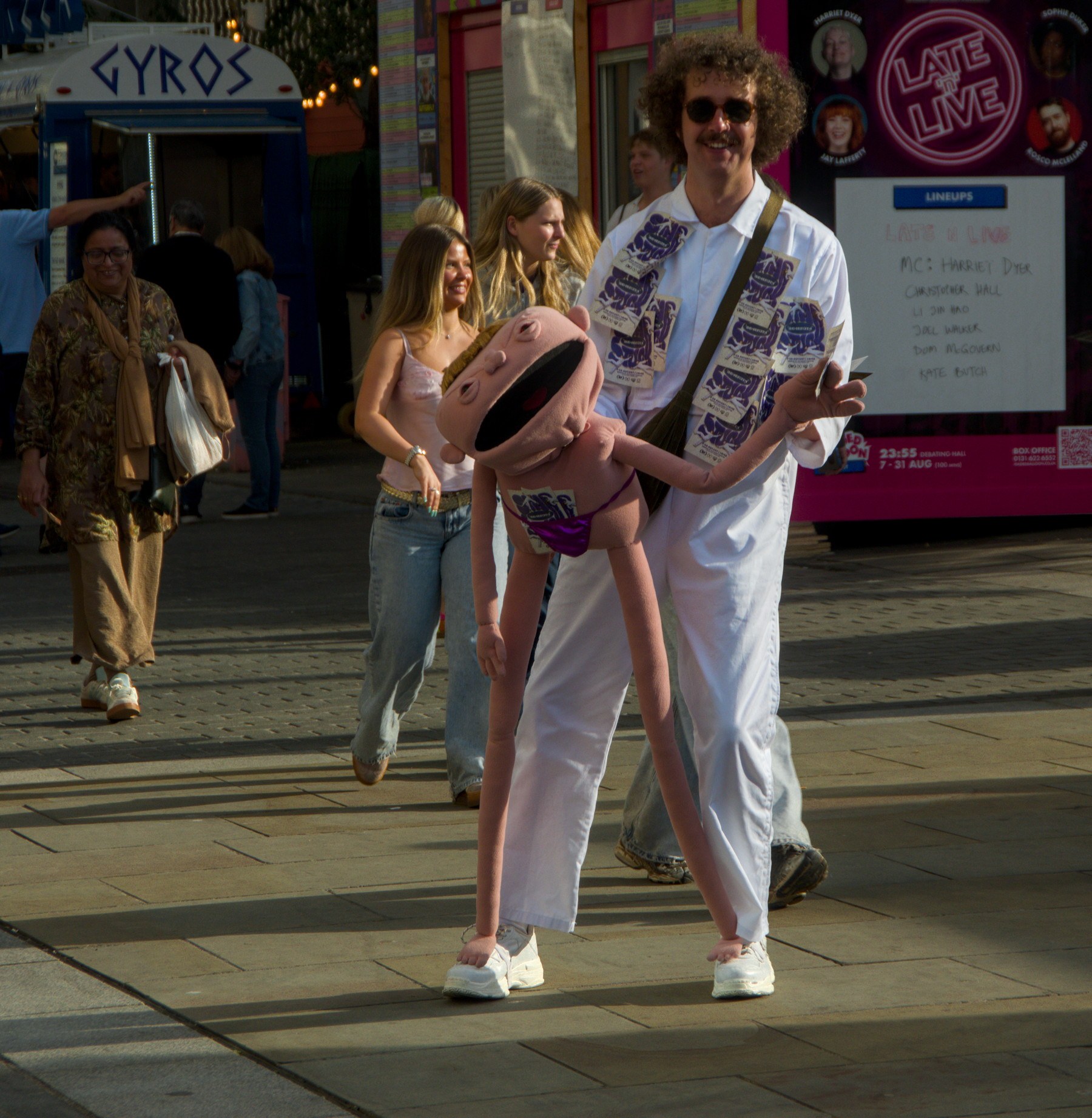 Man in a white outfit carries a large pink cartoon character puppet along an Edinburgh city-centre pavement as pedestrians pass behind him.