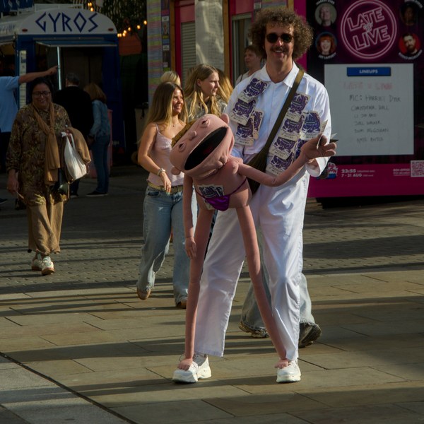 Man in a white outfit carries a large pink cartoon character puppet along an Edinburgh city-centre pavement as pedestrians pass behind him.