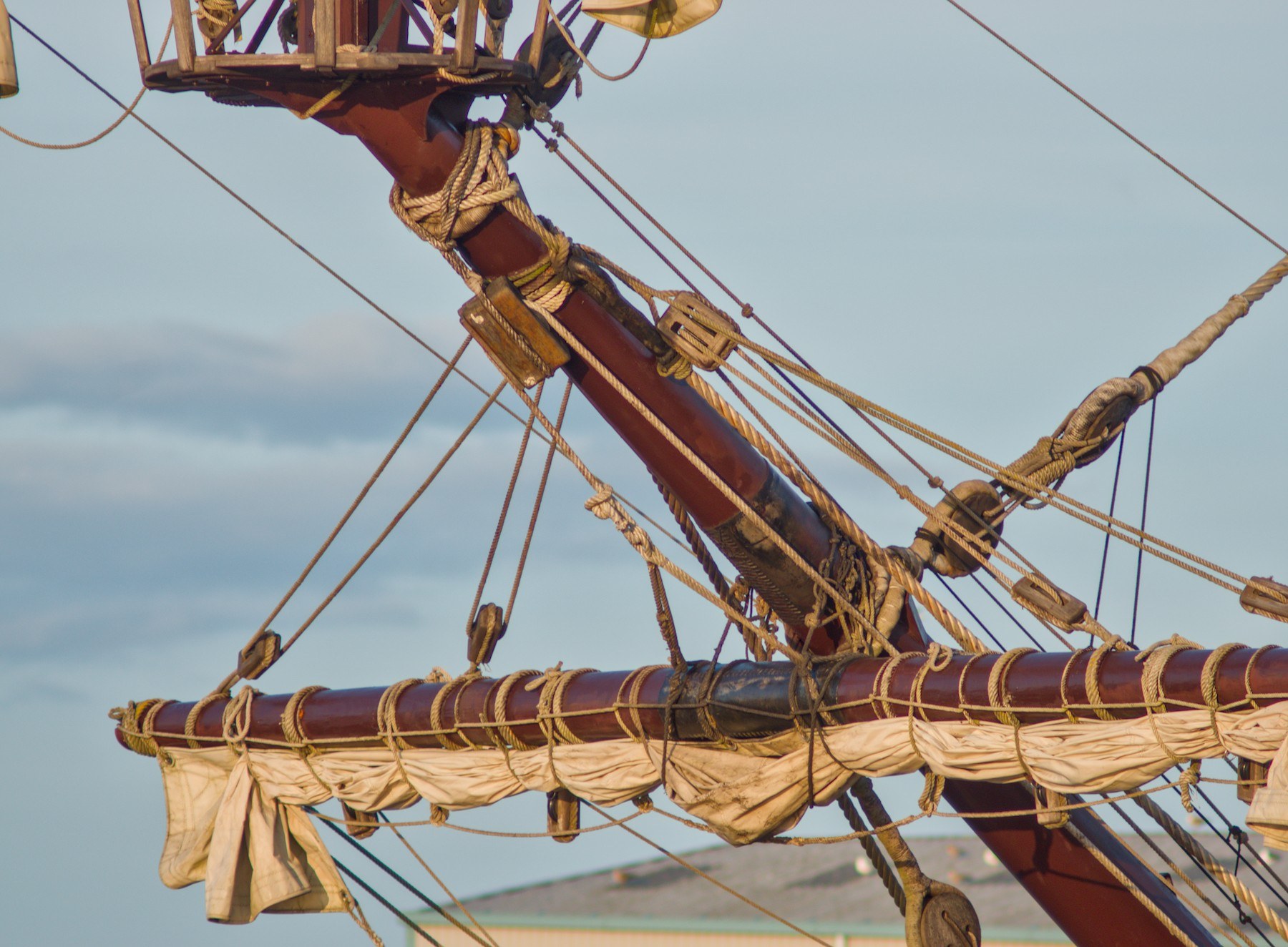 Furled cream sail tied beneath a dark wooden yard, with dense rigging and pulley blocks leading to the angled mast of Galeón Andalucía.
