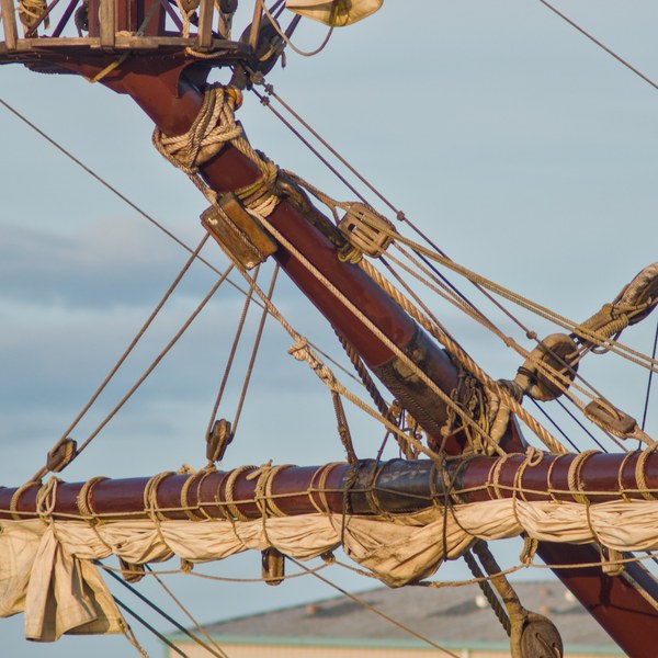 Furled cream sail tied beneath a dark wooden yard, with dense rigging and pulley blocks leading to the angled mast of Galeón Andalucía.