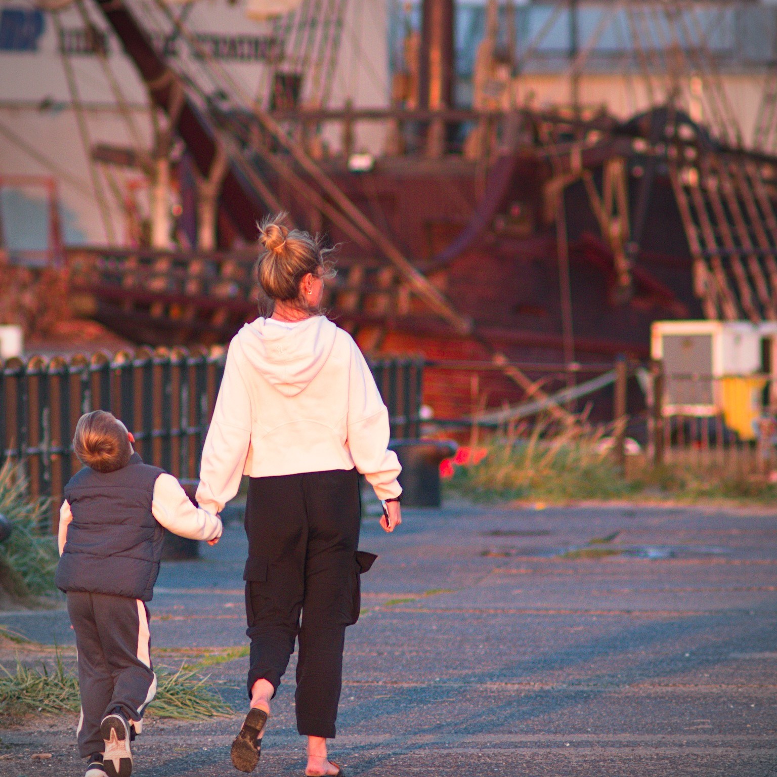 An adult in a white hoodie holds a child’s hand while walking toward the fenced quayside, with Galeón Andalucía’s wooden hull behind them.
