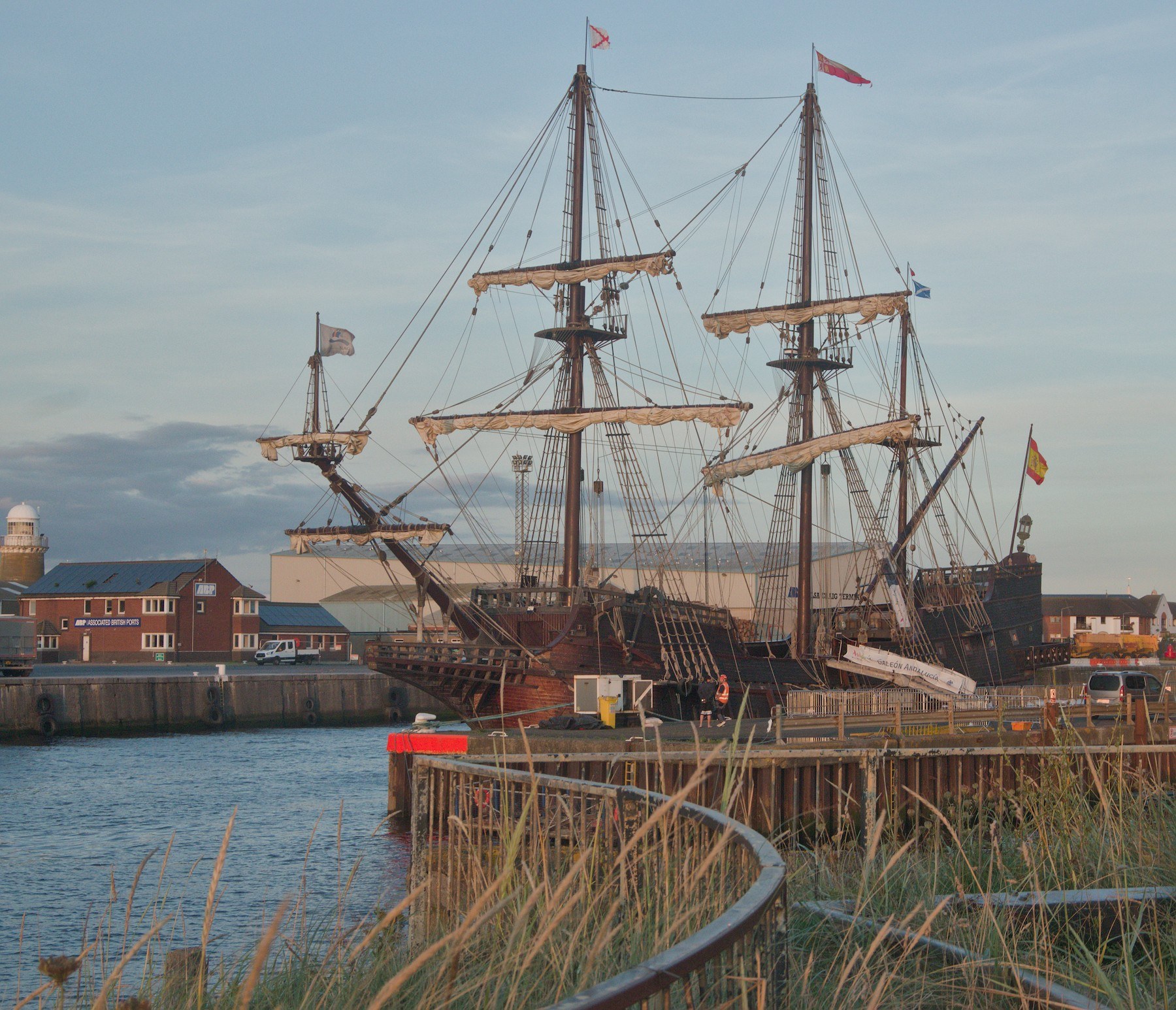 Galeón Andalucía moored at the Port of Ayr, with three tall rigged masts, harbour buildings and a curved rail beside grasses in the foreground.