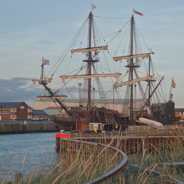Galeón Andalucía moored at the Port of Ayr, with three tall rigged masts, harbour buildings and a curved rail beside grasses in the foreground.
