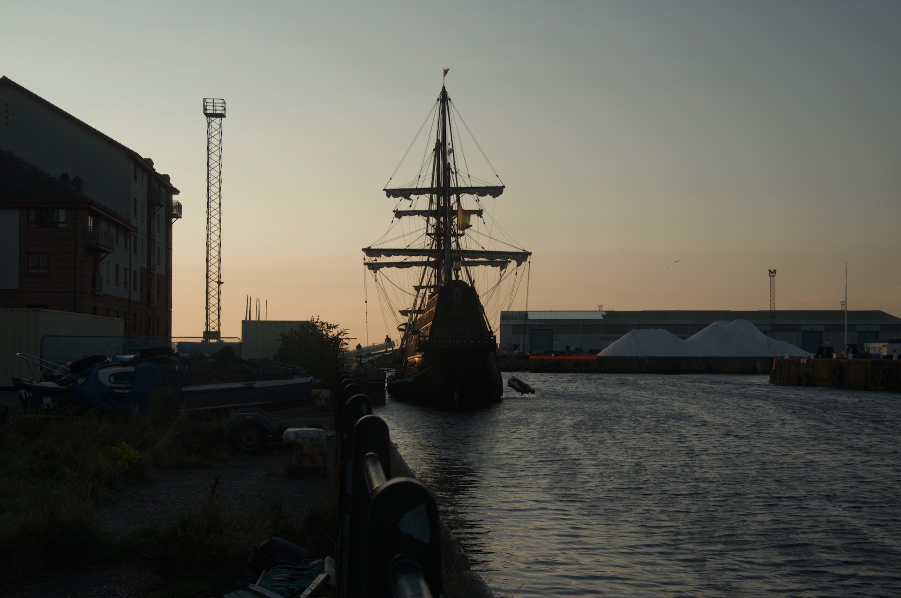 Galeón Andalucía moored in the Port of Ayr channel, silhouetted against an evening sky, with warehouses, a floodlight tower and quay-side railings nearby.