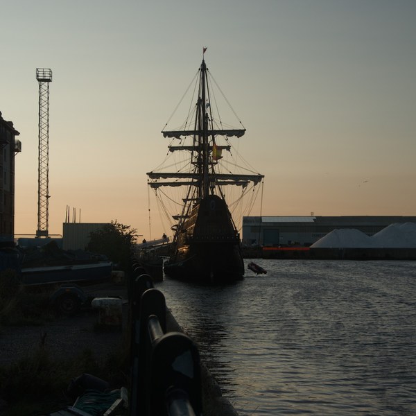 Galeón Andalucía moored in the Port of Ayr channel, silhouetted against an evening sky, with warehouses, a floodlight tower and quay-side railings nearby.
