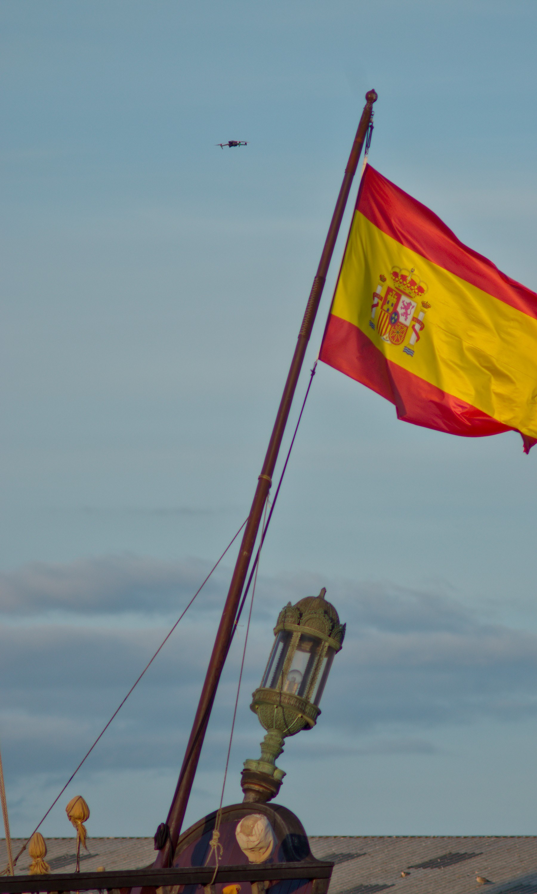 Spanish flag flying from Galeón Andalucía’s mast above an ornate stern lantern, with a small drone in the blue-grey sky.
