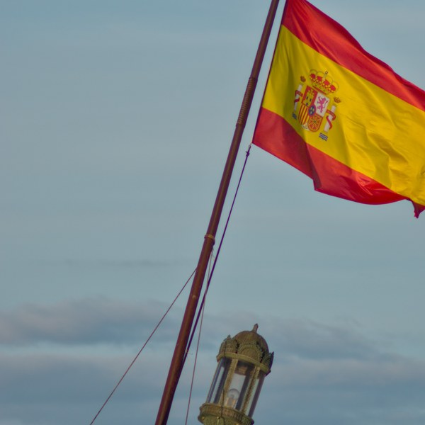Spanish flag flying from Galeón Andalucía’s mast above an ornate stern lantern, with a small drone in the blue-grey sky.