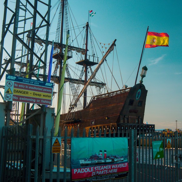 Galeón Andalucía moored behind a metal security gate at the Port of Ayr, with warning signs, a Paddle Steamer Waverley banner, tall ship lines and a Spanish flag against blue sky.