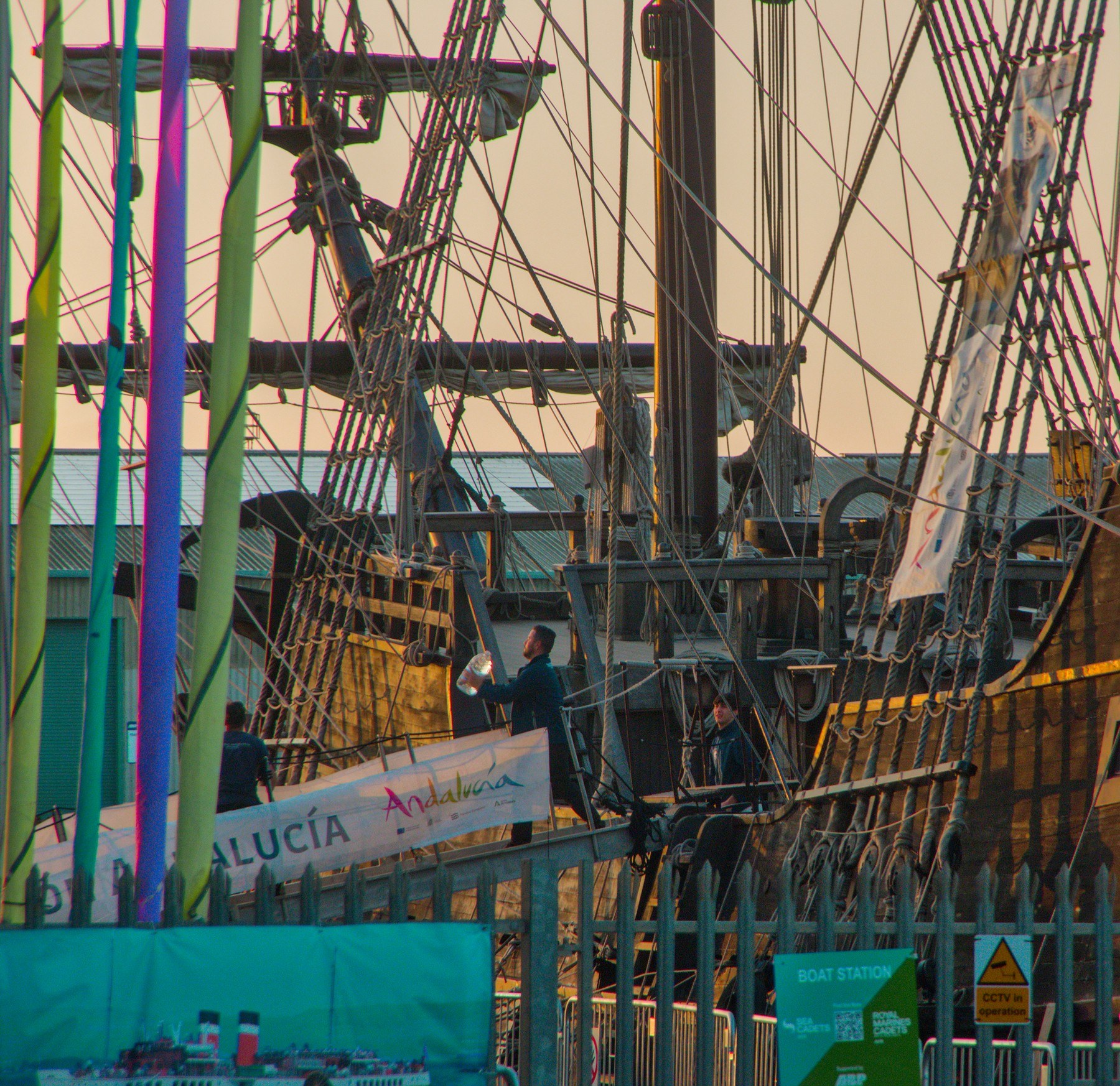 A person on the Galeón Andalucía gangway, framed by green and blue hanging strips, dense rope ladders, masts and the ship’s wooden hull at Port of Ayr.