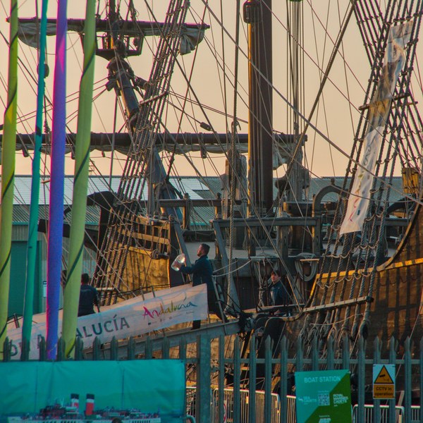 A person on the Galeón Andalucía gangway, framed by green and blue hanging strips, dense rope ladders, masts and the ship’s wooden hull at Port of Ayr.