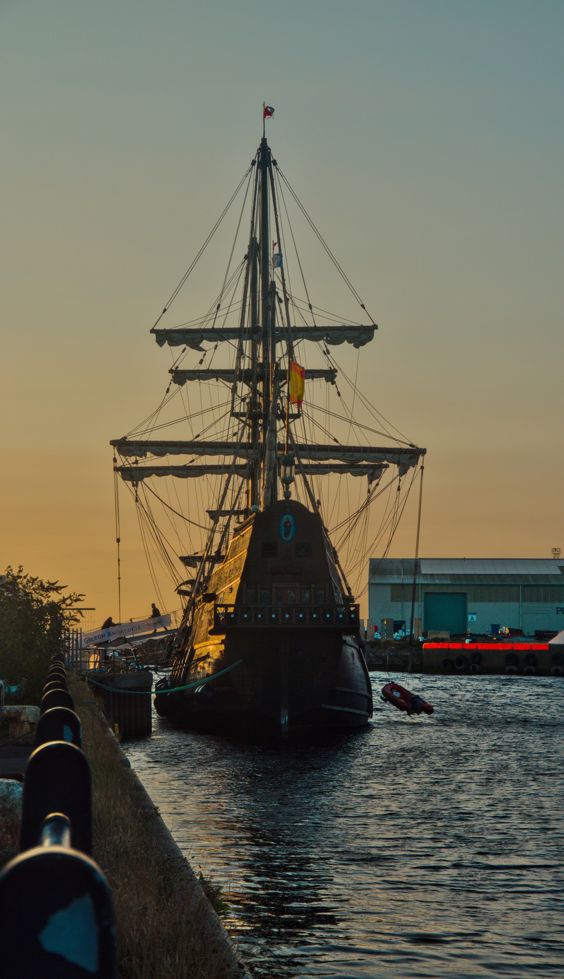 Stern view of Galeón Andalucía moored at Port Of Ayr at sunset, with a Spanish flag, tall yards and ropes above dark water; a small boat floats beside it.
