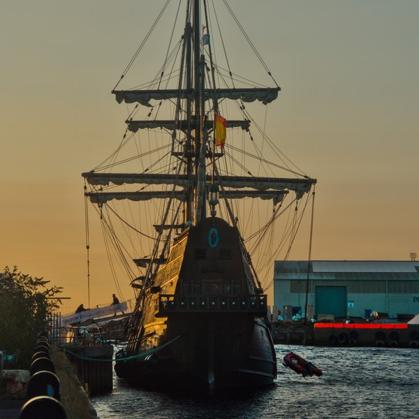 Stern view of Galeón Andalucía moored at Port Of Ayr at sunset, with a Spanish flag, tall yards and ropes above dark water; a small boat floats beside it.