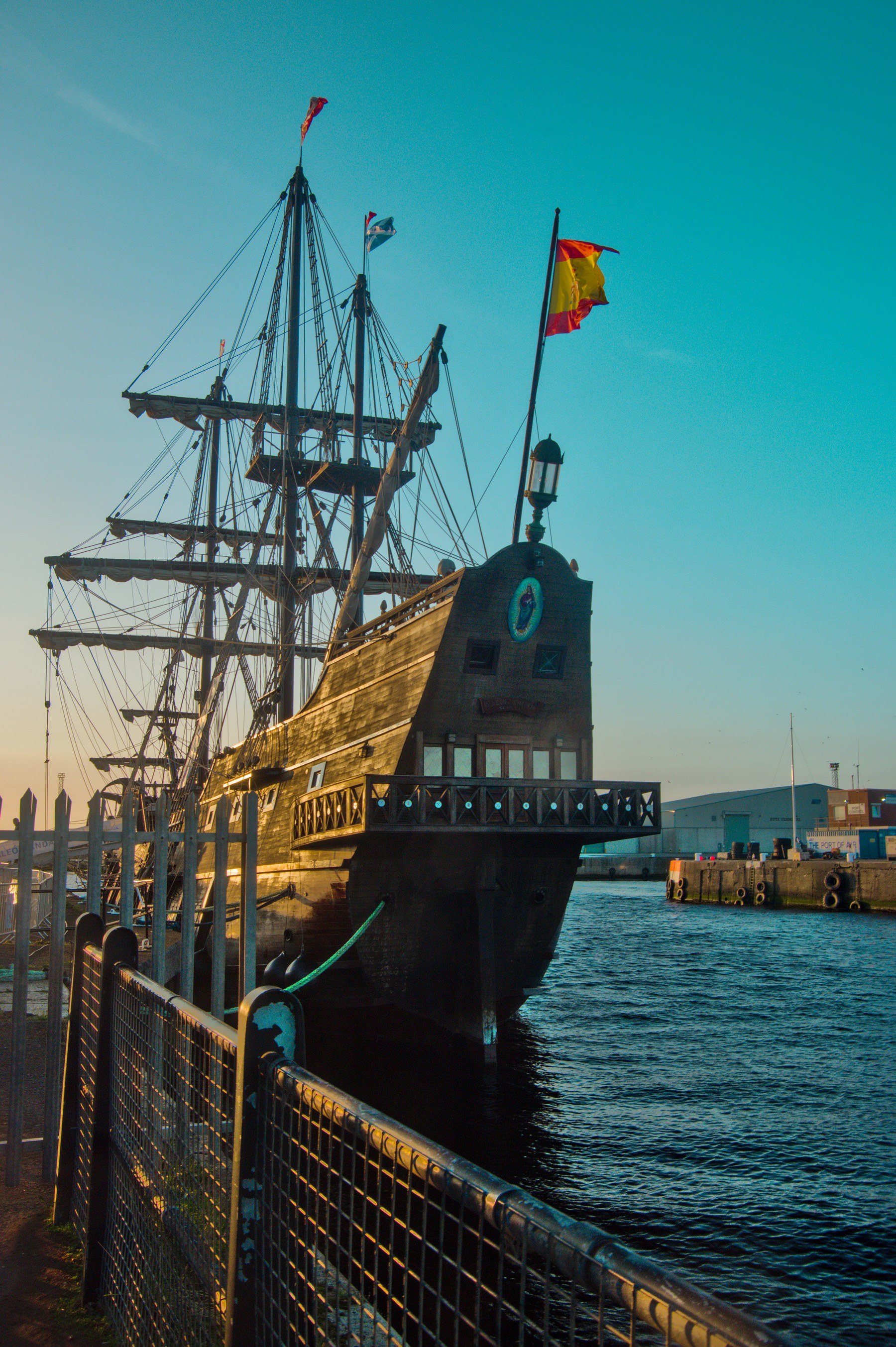 Galeón Andalucía moored at Port of Ayr, seen from behind a metal railing with its wooden rear gallery, tall spars, dense rigging and a Spanish flag against a clear sky.