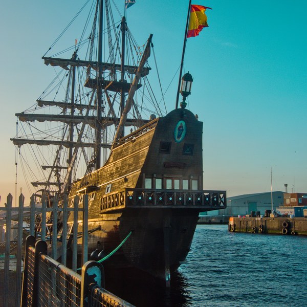 Galeón Andalucía moored at Port of Ayr, seen from behind a metal railing with its wooden rear gallery, tall spars, dense rigging and a Spanish flag against a clear sky.