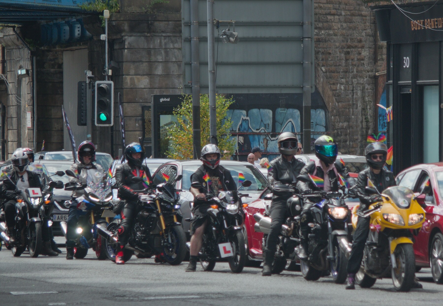 A group of helmeted motorcyclists, several carrying rainbow flags, ride along a city street; some bikes display L plates.
