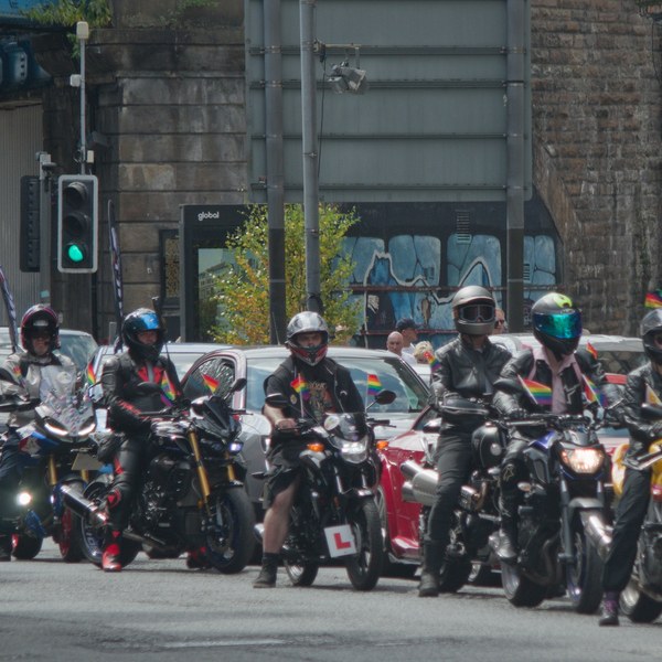 A group of helmeted motorcyclists, several carrying rainbow flags, ride along a city street; some bikes display L plates.