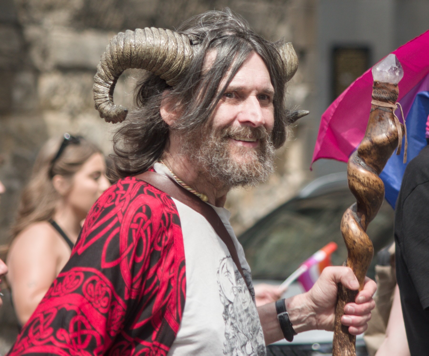 Smiling bearded person wearing a large ram-horn headdress and red patterned garment, holding a carved wooden staff topped with a crystal; colourful flags and other attendees appear behind them.