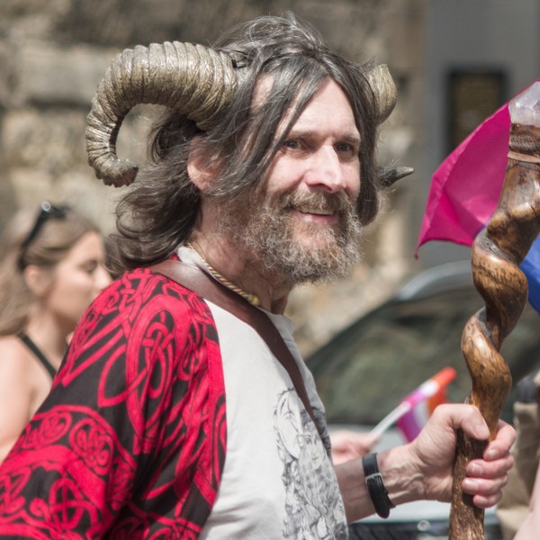 Smiling bearded person wearing a large ram-horn headdress and red patterned garment, holding a carved wooden staff topped with a crystal; colourful flags and other attendees appear behind them.