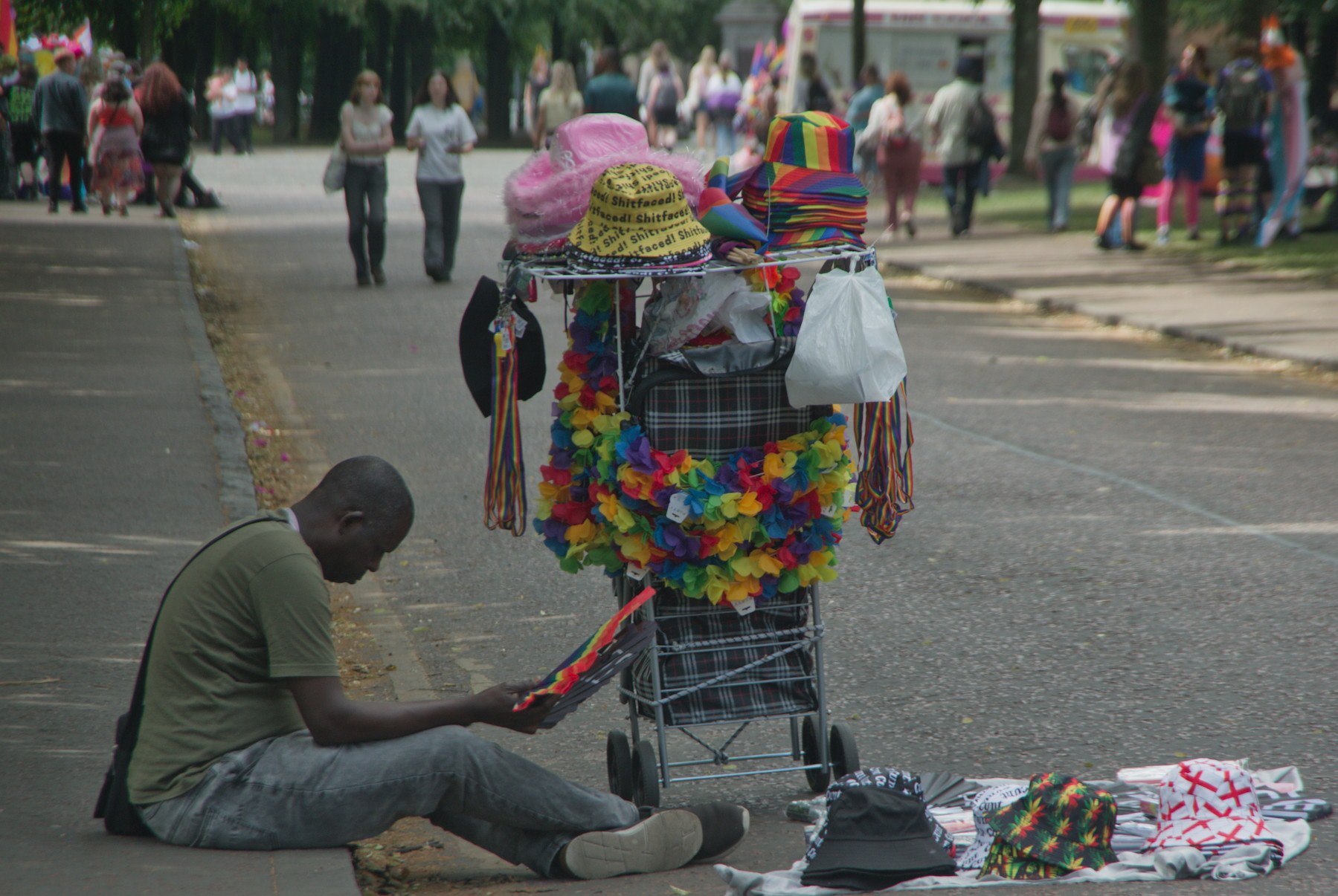 A seated man beside a wheeled cart covered with rainbow leis, hats and colourful accessories, with more merchandise spread on the pavement at Glasgow Green.