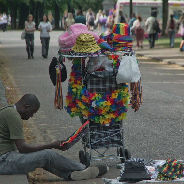 A seated man beside a wheeled cart covered with rainbow leis, hats and colourful accessories, with more merchandise spread on the pavement at Glasgow Green.