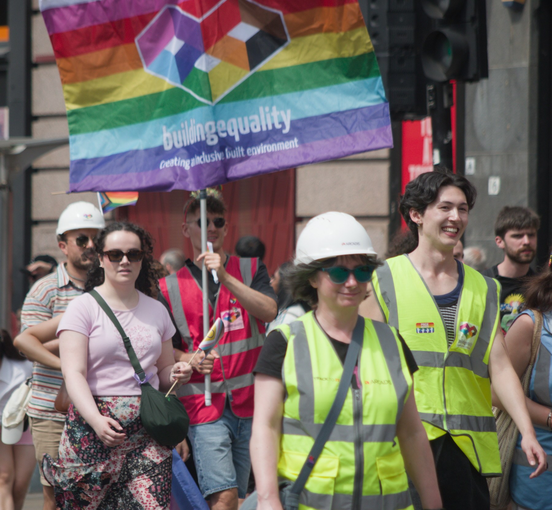 Pride marchers in high-visibility vests and hard hats walk beneath a rainbow “Building Equality” banner.