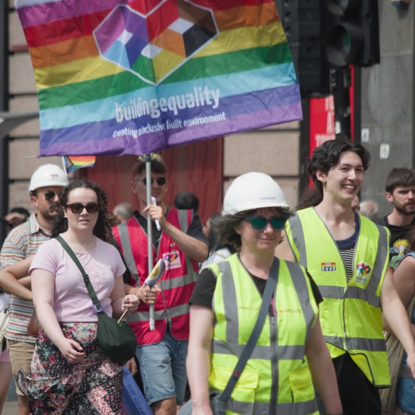 Pride marchers in high-visibility vests and hard hats walk beneath a rainbow “Building Equality” banner.
