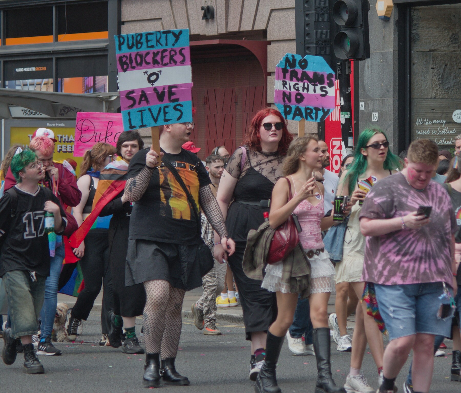 Pride marchers on a city street, including a person holding a sign reading “Puberty Blockers Save Lives” and another sign reading “No Trans Rights No Pride.”