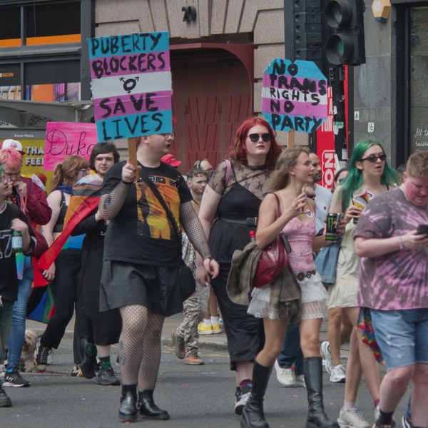 Pride marchers on a city street, including a person holding a sign reading “Puberty Blockers Save Lives” and another sign reading “No Trans Rights No Pride.”