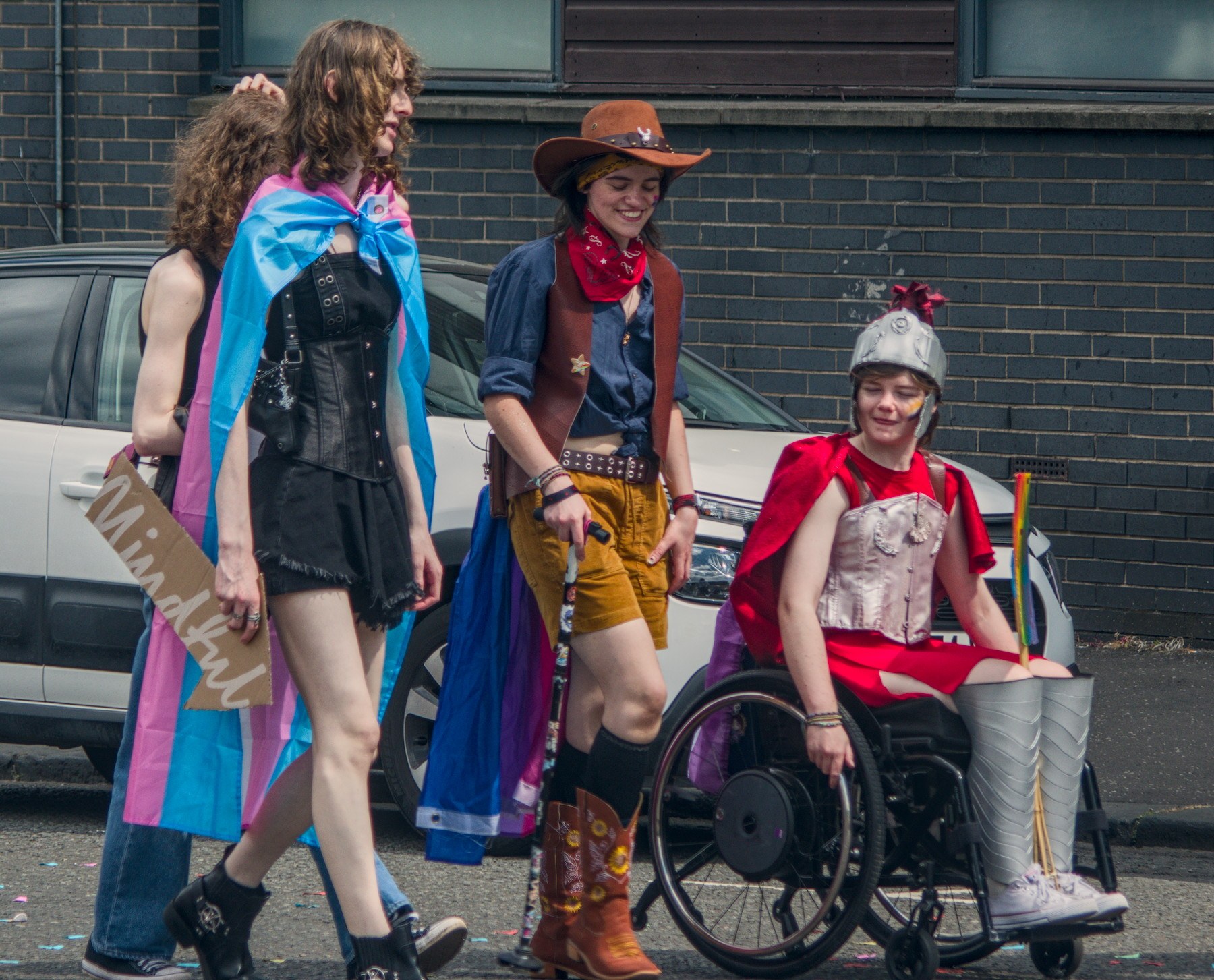 Three Pride participants walk beside parked cars: one wears a transgender flag as a cape, one is dressed in cowboy-inspired clothing, and one uses a wheelchair while wearing a red cape and silver costume armour.