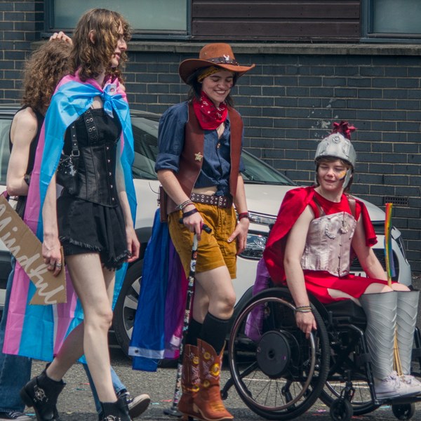 Three Pride participants walk beside parked cars: one wears a transgender flag as a cape, one is dressed in cowboy-inspired clothing, and one uses a wheelchair while wearing a red cape and silver costume armour.