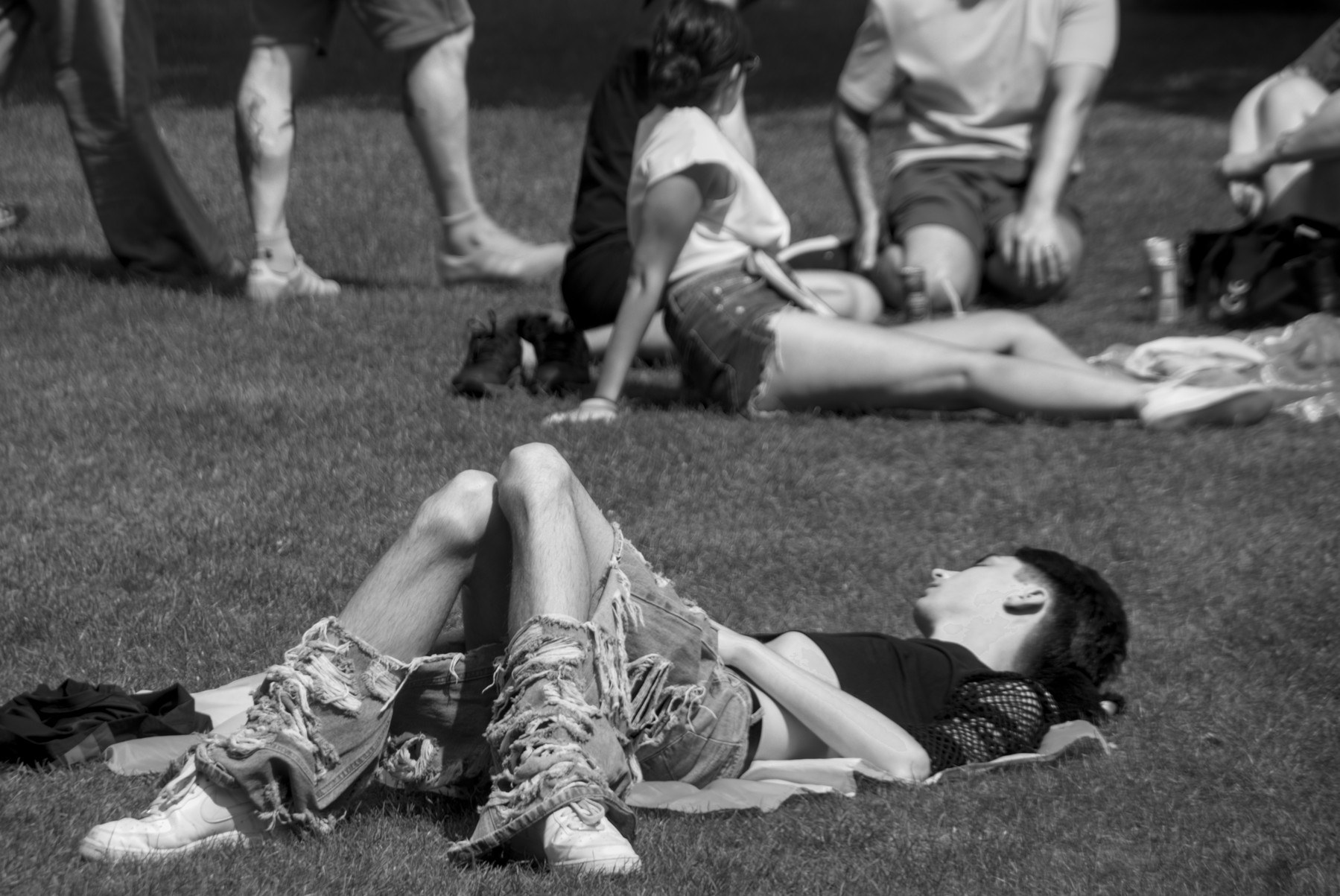 Black-and-white photograph of a person resting on grass in fringed boots and sunglasses, with other seated people behind them.